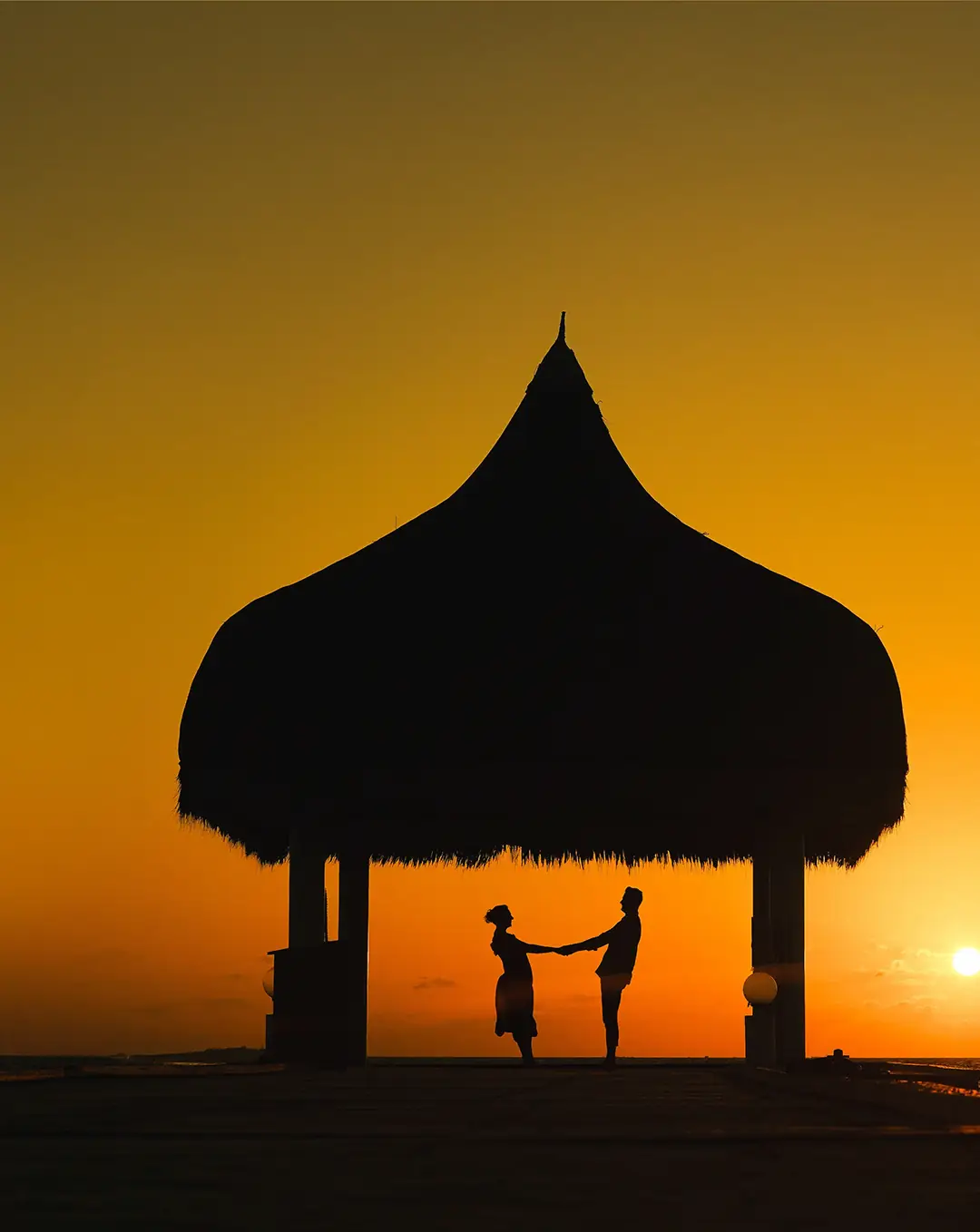 A couple holding hands under a thatched gazebo at sunset